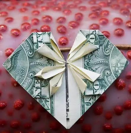A crisp US dollar bill folded into a small heart shape held in a person's hand.